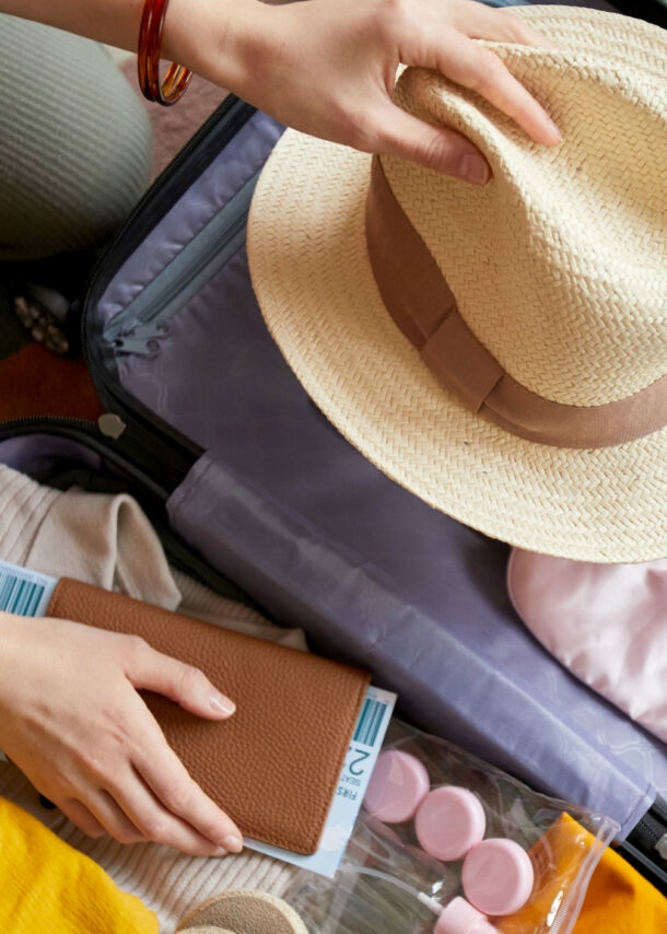 Aufsicht von zwei Händen mit Strohhut und Reiseunterlagen über einem geöffneten, gepackten Koffer mit Reiseutensilien.