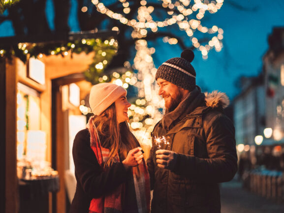 Eine Frau und ein Mann stehen auf dem Weihnachtsmarkt und schauen sich an, in ihren Händen halten sie Wunderkerzen