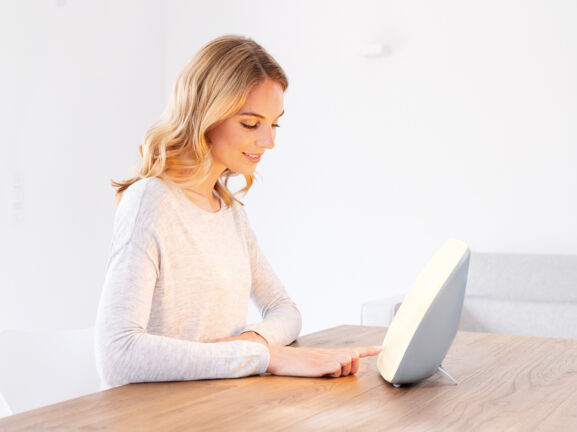 Eine junge Frau sitzt an einem Tisch vor einer Tageslichtlampe