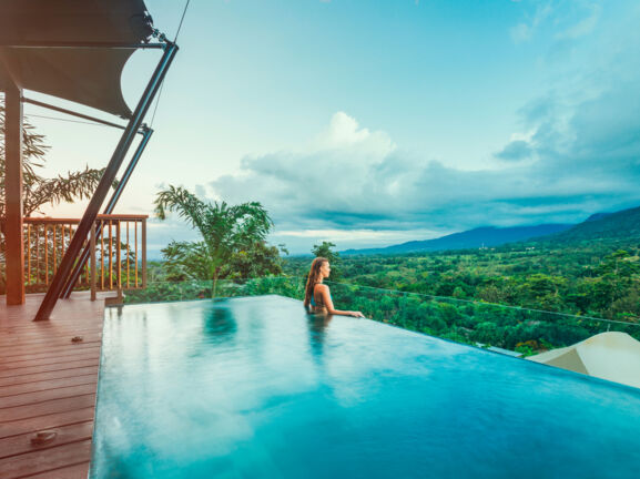 Profilansicht einer Frau, die aus einem Infinity-Pool über eine tropische Hügellandschaft schaut