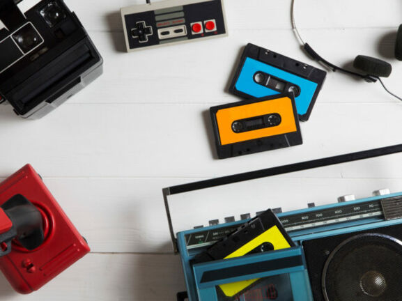 Auf einem Tisch liegen mehrere analoge Medien in Form von Kassetten, einer Polaroidkamera, einem Kassettenrekorder, einem NES-Gamepad und Joystick