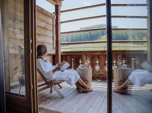 Eine Frau in weißem Bademantel sitzt mit einem Buch auf einer Terrasse eines Holzchalets mit Blick in die Berge