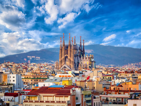 Stadtpanorama von Barcelona mit der emporragenden Sagrada Familia vor einem Gebirgszug im Hintergrund.