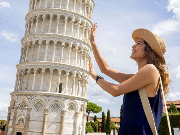 Profilansicht einer jungen Frau mit Strohhut, die aus der fotografischen Perspektive den Schiefen Turm von Pisa mit ihren Händen zu halten scheint.