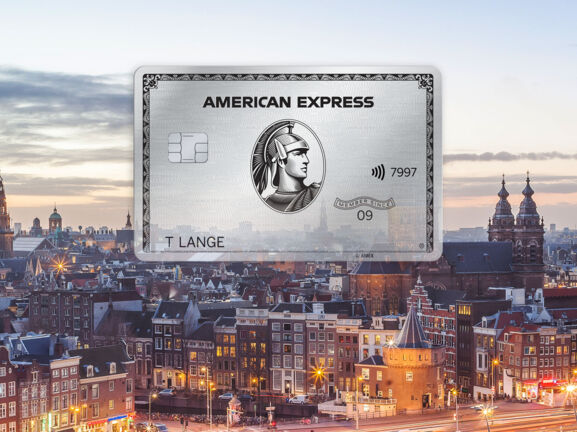 Fotocollage mit Freisteller einer silbernen Kreditkarte von American Express vor dem Stadtpanorama von Amsterdam in der Abenddämmerung.