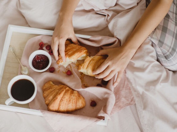 Auf einem Bett liegt ein Tablett mit Croissants, einer Tasse Kaffee, ein Schälchen Marmelade, einem Löffel und Früchten. Dahinter sitzt eine unkenntliche Person, die eines der Croissants auseinanderreißt.