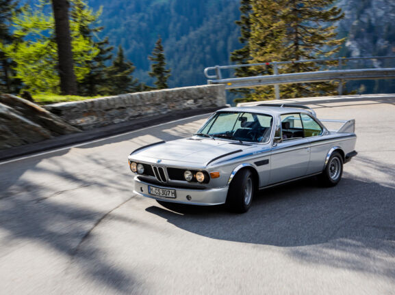 Ein fahrender silberner BMW 3.0 CSL in einer Kurve auf einer Straße in den Bergen.