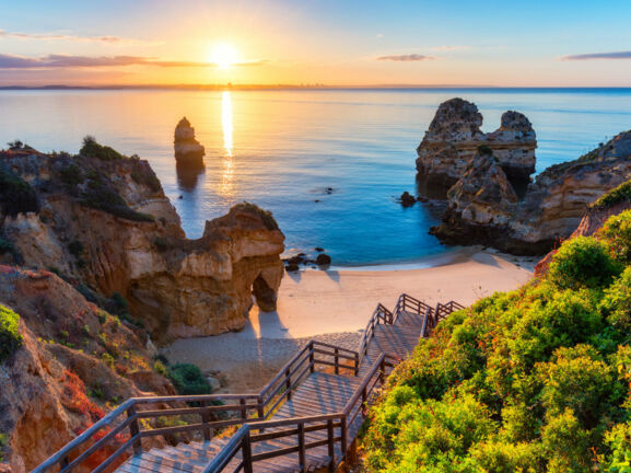 Der Strand Praia do Camilo in Portugal bei Sonnenuntergang, zu dem über Felsen eine Holztreppe nach unten führt.