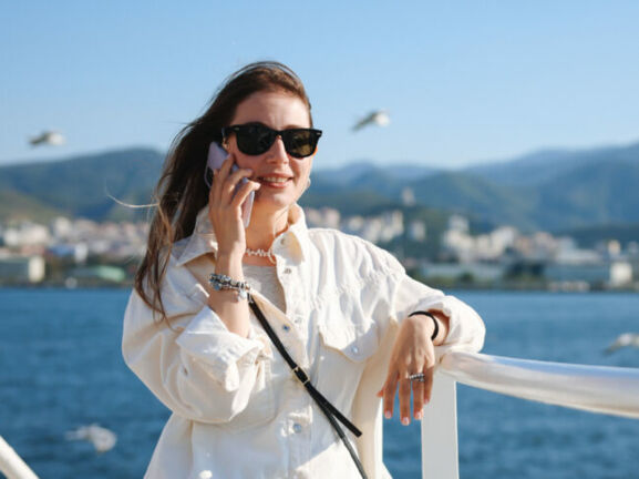 Eine Frau mit Sonnenbrille und wehenden Haaren telefoniert mit einem Smartphone an der Reling eines Schiffes auf dem Meer, im Hintergrund fliegen Möwen vor Stadtpanorama an einer Küste.